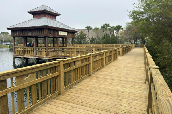 Wooden walkway on a lake surrounded by trees, with a covered pavilion to the left.