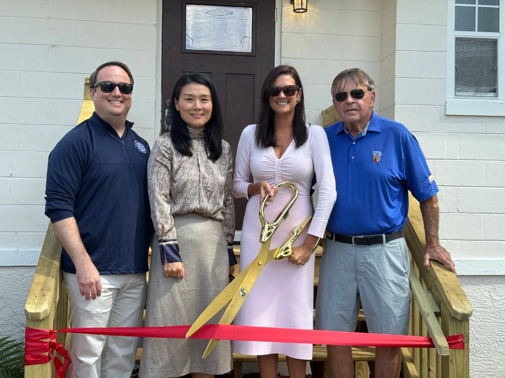 Four people stand on the wooden steps of a house behind a red ribbon, while one woman prepares to cut the ribbon with a huge pair of golden scissors.