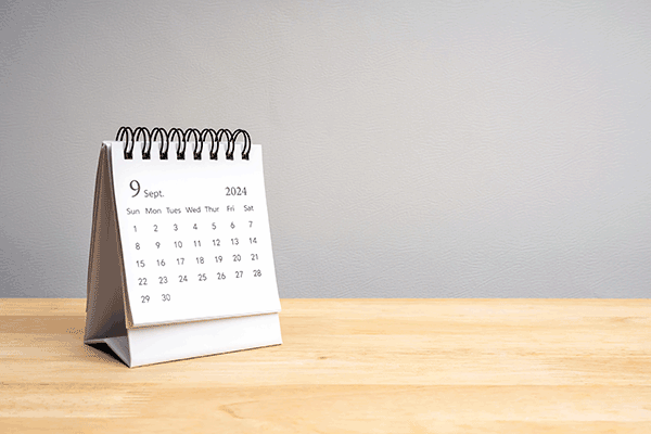 A white desk calendar open to September on a light wood desk with a gray wall behind it.