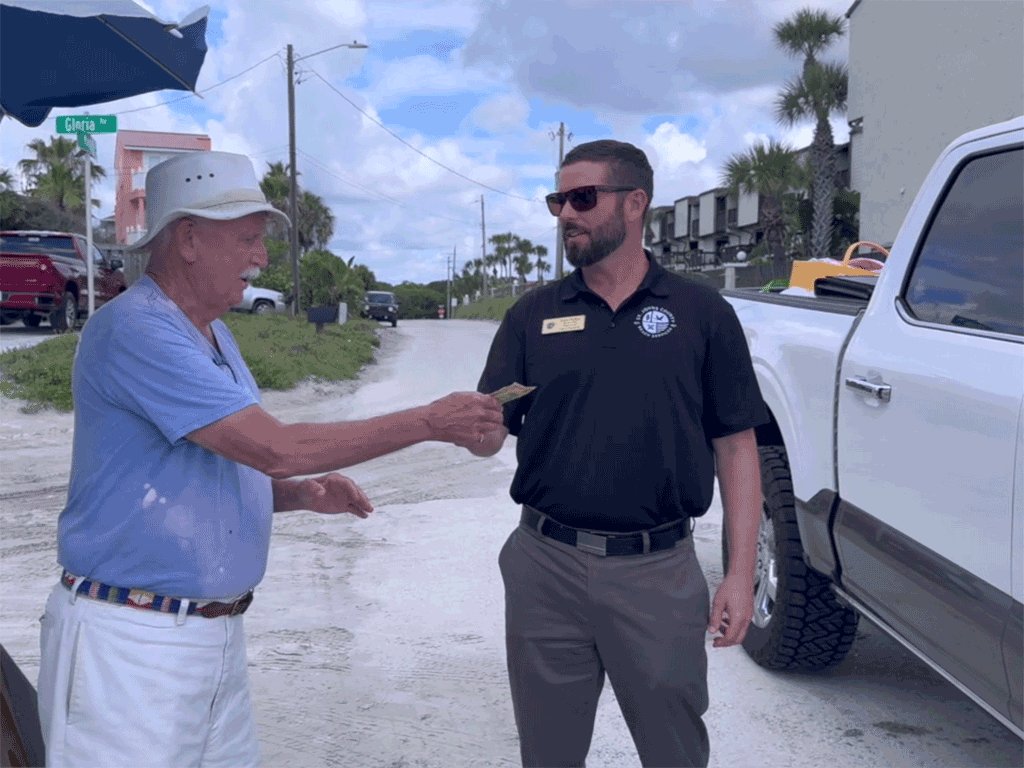 A man in sunglasses passes a dollar bill from the driver of a pickup truck to an older man in a hat on a sandy road.