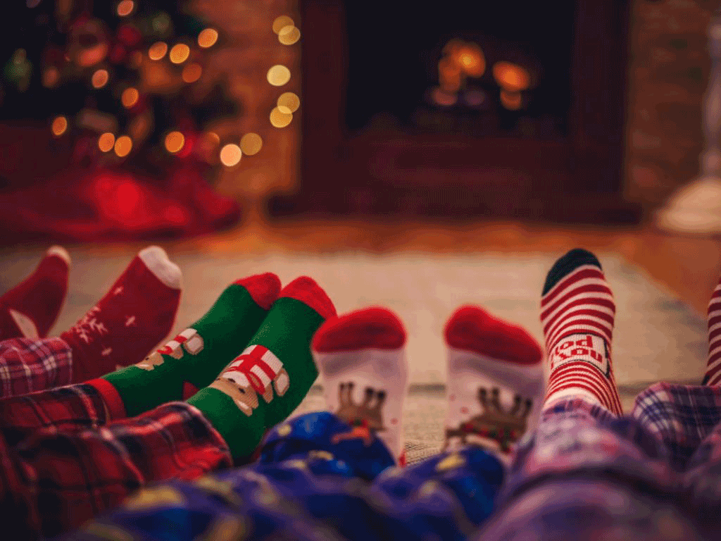 Four pairs of legs and feet in festive pajamas and socks, with a Christmas tree and fireplace in a decorated living room the background