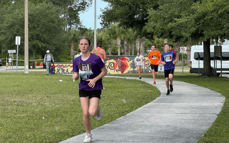 A line of kids in running outfits and racing bibs sprints down a sidewalk during a triathlon.