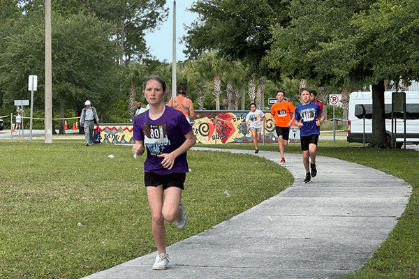 A line of kids in running outfits and racing bibs sprints down a sidewalk during a triathlon.